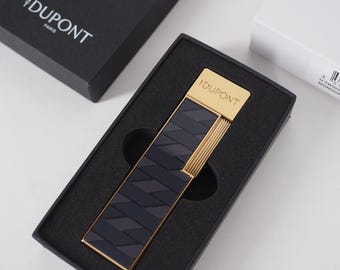 May include: A gold-toned lighter with a navy blue and black geometric pattern, presented in a black box. The lighter has the "DUPONT PARIS" logo. The box is open, revealing the lighter nestled in black foam.