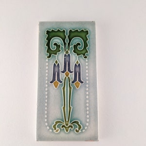 May include: A vintage ceramic tile featuring a blue, green and yellow floral design. The tile has a light blue background and a white border with a dotted pattern.