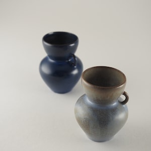 Può includere: Due vasi in ceramica, uno blu e uno con una smaltatura blu e marrone screziata. Entrambi i vasi hanno un corpo arrotondato e un piccolo manico.