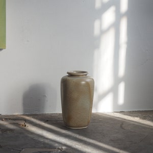 May include: A light brown ceramic vase with a textured surface sits on a grey concrete floor with cracks. The vase is in the centre of the image and is illuminated by sunlight streaming through a window.