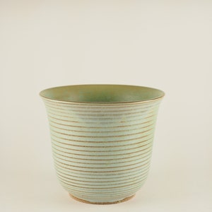 May include: A light green ceramic planter with a textured, striped design. The planter has a wide, round opening and a slightly flared lip.