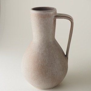 May include: A light brown ceramic vase with a rounded body and a single handle. The vase has a textured surface and a slightly off-white colour.