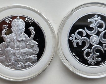 Könnte beinhalten: Eine Silbermünze mit einer Darstellung der hinduistischen Gottheit Ganesha auf der einen Seite und dem Om-Symbol auf der anderen. Die Münze befindet sich in einem durchsichtigen Plastiketui.
