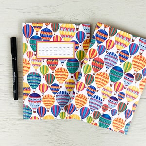 May include: Two notebooks with a white background and colourful hot air balloon designs. The notebooks have a blank space for writing on the front cover.