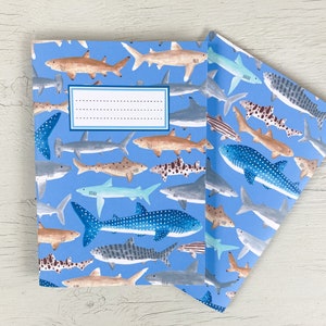 May include: A blue notebook and a matching folded paper, both covered in a pattern of various shark illustrations. The sharks are in shades of brown, gray, blue, and white, set against a light blue background. The notebook has a white label.