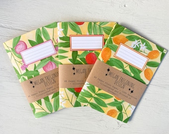May include: Three small notebooks with illustrated covers featuring peach, citrus fruit, and floral designs. Each notebook has a brown paper label with the text "MEGAN TREGONING DESIGN" and details about the paper and printing.