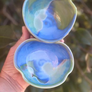 May include: Two handmade ceramic bowls with a unique, organic shape. The bowls feature a blend of blue, green, and white glaze, creating a marbled effect. The rims are a lighter green, and the interiors are a vibrant blue.