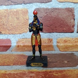 Puede incluir: Una figura negra del dios egipcio Thot, sosteniendo un bastón con un orbe rojo en la parte superior. La figura lleva un tocado dorado y azul y una falda dorada. La base de la figura es negra y tiene la palabra "Thoth" escrita en ella.