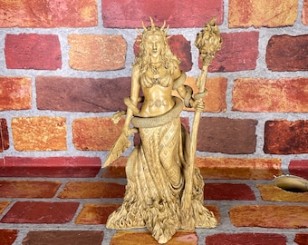 May include: A golden statue of a woman with a crown of thorns and a snake wrapped around her arm. She is holding a torch in her right hand and a sword in her left hand. The statue is standing on a brick wall.