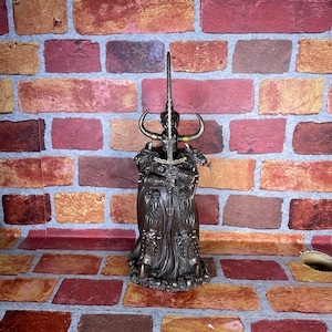 May include: A dark bronze figurine of a warrior with a sword and horns, standing on a brick background.