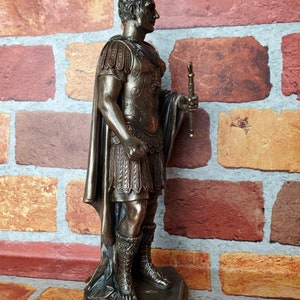 Puede incluir: Estatua de bronce de un emperador romano, de pie sobre una base rocosa. Lleva una toga y una armadura, y sostiene un bast&oacute;n en su mano derecha.
