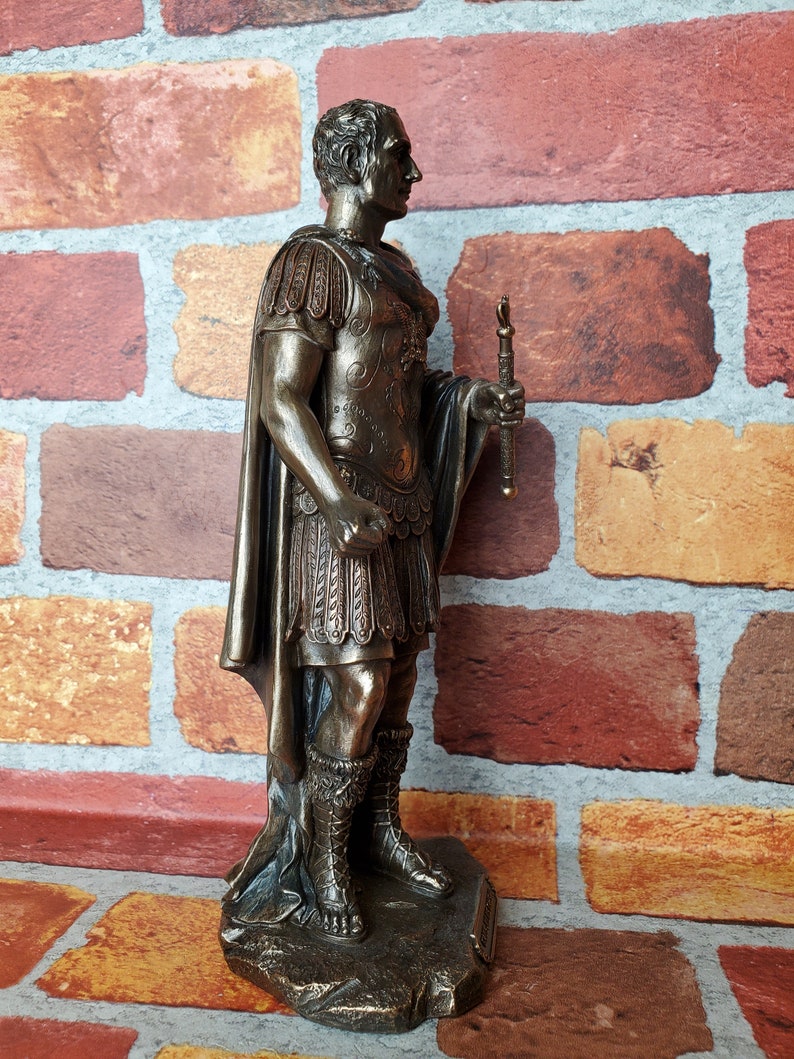 Puede incluir: Estatua de bronce de un emperador romano, de pie sobre una base rocosa. Lleva una toga y una armadura, y sostiene un bast&oacute;n en su mano derecha.
