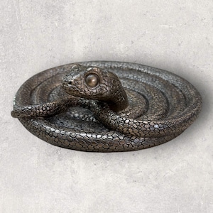 Peut inclure: Une sculpture en bronze d'un serpent enroulé en cercle. Le serpent a une surface texturée détaillée et un œil réaliste.