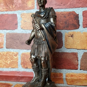 Puede incluir: Una estatua de bronce de Julio C&eacute;sar, de pie con un cetro en su mano derecha. La estatua est&aacute; sobre una base con la inscripci&oacute;n "GAIUS JULIUS CAESAR".