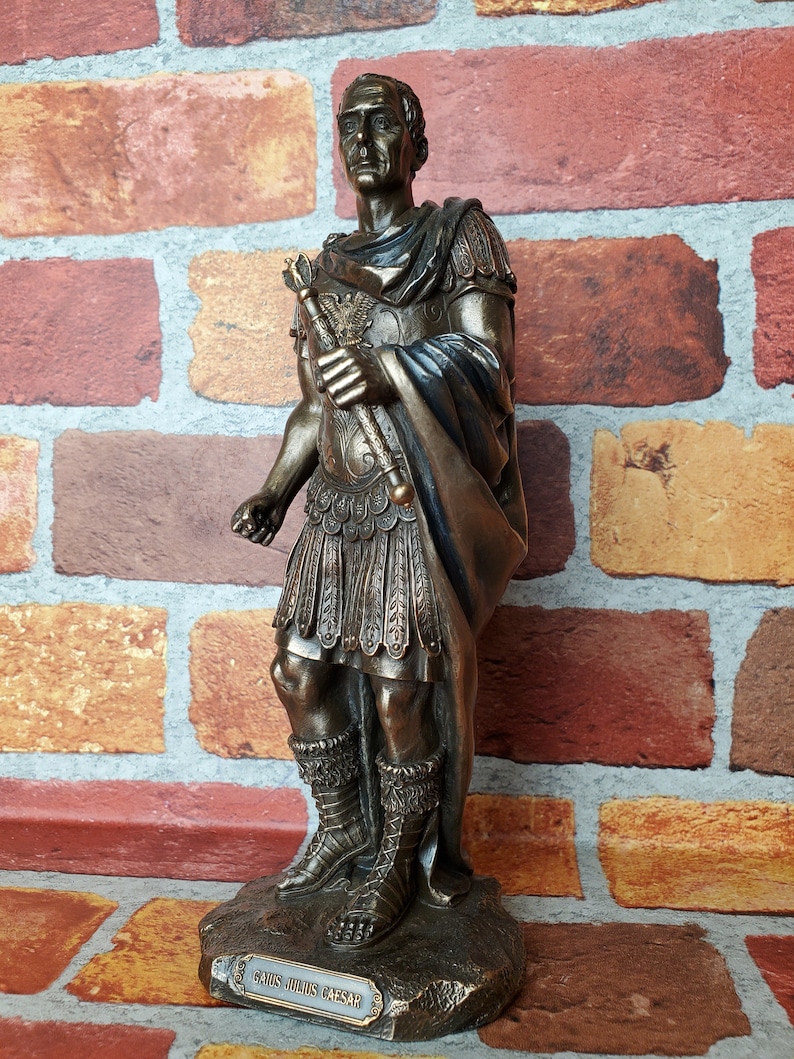 Puede incluir: Una estatua de bronce de Julio C&eacute;sar, de pie con un cetro en su mano derecha. La estatua est&aacute; sobre una base con la inscripci&oacute;n "GAIUS JULIUS CAESAR".