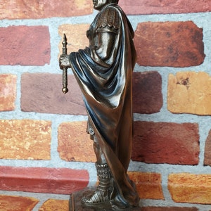 Puede incluir: Estatua de bronce de un soldado romano con toga y sosteniendo una lanza. La estatua est&aacute; de pie sobre una base rocosa.