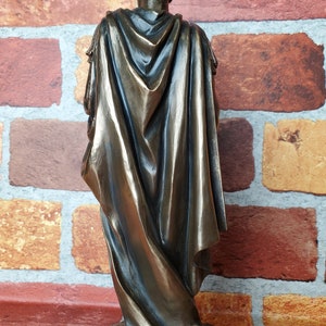 Puede incluir: Una estatua de bronce de una persona que lleva una toga y una capa. La estatua est&aacute; de pie sobre una base y est&aacute; de espaldas a la c&aacute;mara.