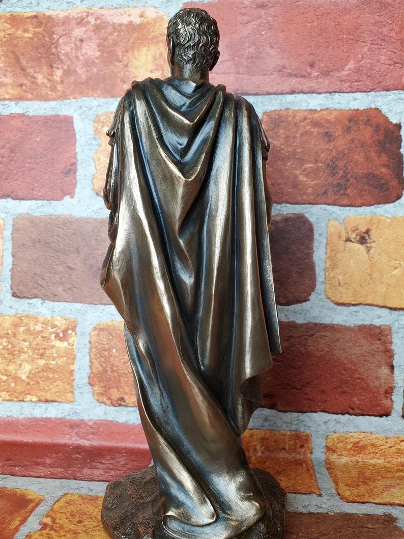 Puede incluir: Una estatua de bronce de una persona que lleva una toga y una capa. La estatua est&aacute; de pie sobre una base y est&aacute; de espaldas a la c&aacute;mara.