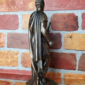 Puede incluir: Estatua de bronce de un emperador romano con una toga y un bast&oacute;n. La estatua est&aacute; sobre una base rocosa.