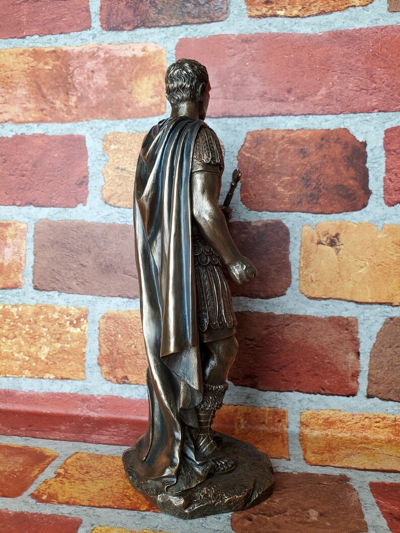 Puede incluir: Estatua de bronce de un emperador romano con una toga y un bast&oacute;n. La estatua est&aacute; sobre una base rocosa.