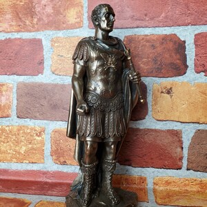 Puede incluir: Estatua de bronce de un emperador romano con toga y armadura, sosteniendo un cetro. La estatua est&aacute; sobre una base con la inscripci&oacute;n "CAESAR AUGUSTUS".