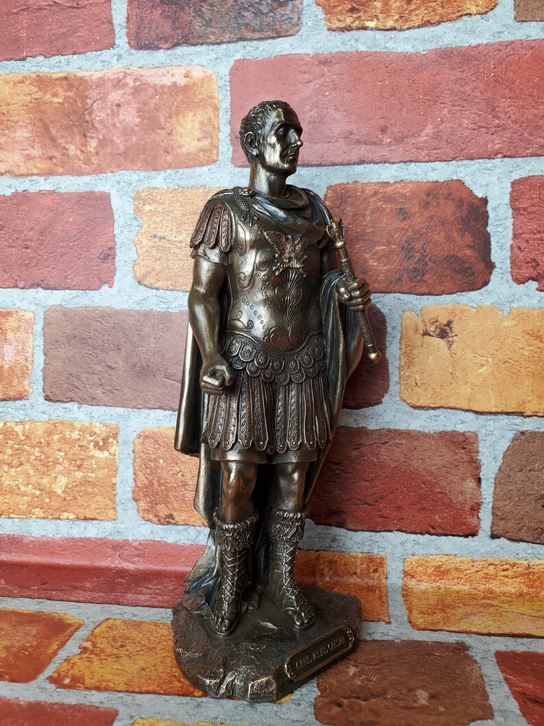 Puede incluir: Estatua de bronce de un emperador romano con toga y armadura, sosteniendo un cetro. La estatua est&aacute; sobre una base con la inscripci&oacute;n "CAESAR AUGUSTUS".