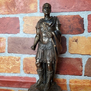 Puede incluir: Estatua de bronce de Julio C&eacute;sar, de pie con una toga y armadura romanas, sosteniendo un bast&oacute;n. La estatua est&aacute; sobre una base con la inscripci&oacute;n "Gaius Julius Caesar".