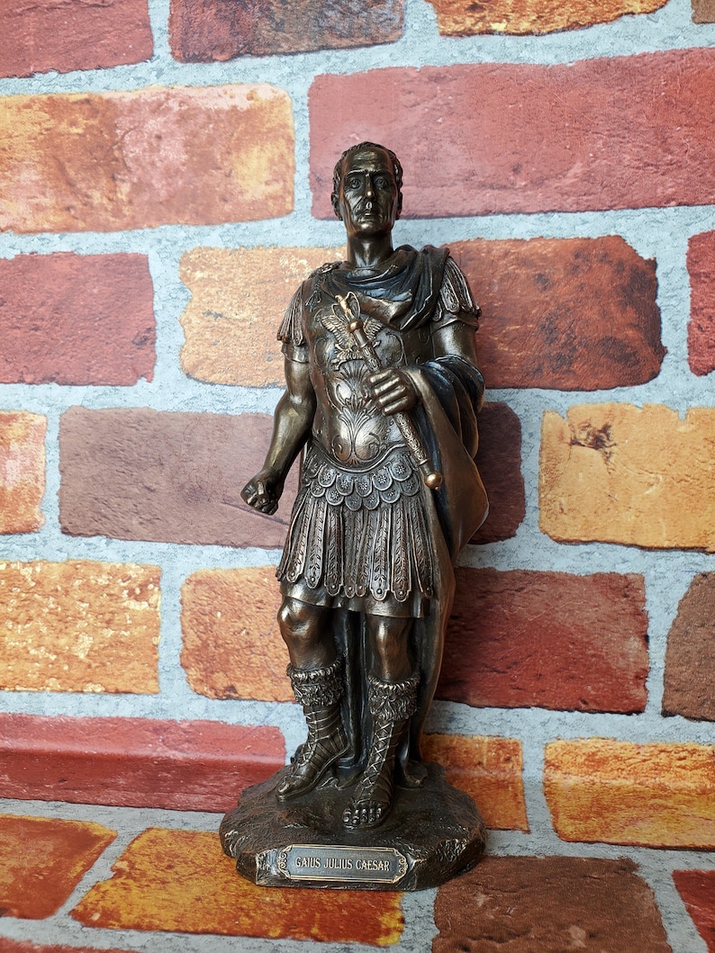 Puede incluir: Estatua de bronce de Julio C&eacute;sar, de pie con una toga y armadura romanas, sosteniendo un bast&oacute;n. La estatua est&aacute; sobre una base con la inscripci&oacute;n "Gaius Julius Caesar".