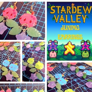 May include: A collection of handmade earrings featuring designs inspired by the video game Stardew Valley. The earrings showcase Junimo characters, stars, and fruit-shaped charms in pink, blue, and green, with green leaf and purple star accents.