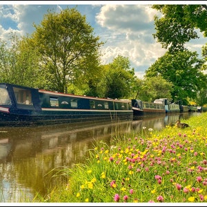 Op de afbeelding: Een rij smalle boten ligt aangemeerd langs een kanaal, met weelderige groene bomen en een blauwe lucht met witte wolken op de achtergrond. De oever van het kanaal is bezaaid met roze en gele wilde bloemen.