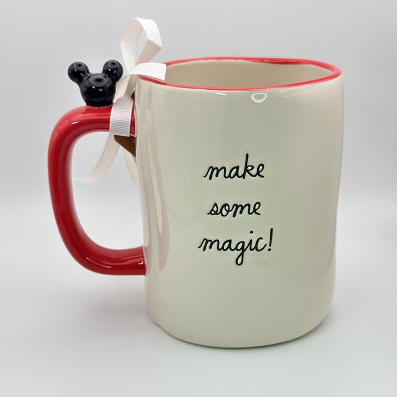 Op de afbeelding: Witte keramische mok met een rode rand en handvat. De mok heeft een zwart Mickey Mouse hoofd bevestigd aan het handvat en de tekst "make some magic!" gedrukt op de zijkant.