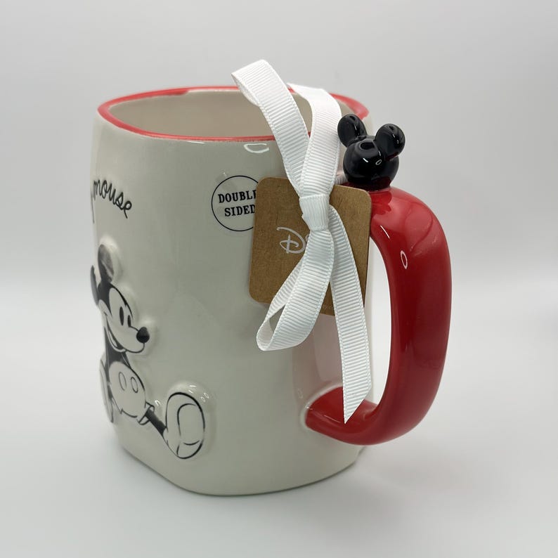 Op de afbeelding: Een witte keramische mok met een rode handgreep en een zwart-witte afbeelding van Mickey Mouse. De mok heeft een rode rand en de woorden "Double Sided" staan ​​gedrukt op de zijkant. De mok is vastgebonden met een wit lint en een bruin label met het woord "Disney" erop gedrukt. Er zit een zwart Mickey Mouse-oor op de handgreep.