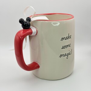Op de afbeelding: Witte keramische mok met een rode handgreep en een rode rand. De mok heeft een zwart Mickey Mouse hoofd op de handgreep en de tekst "make some magic!" aan de zijkant.