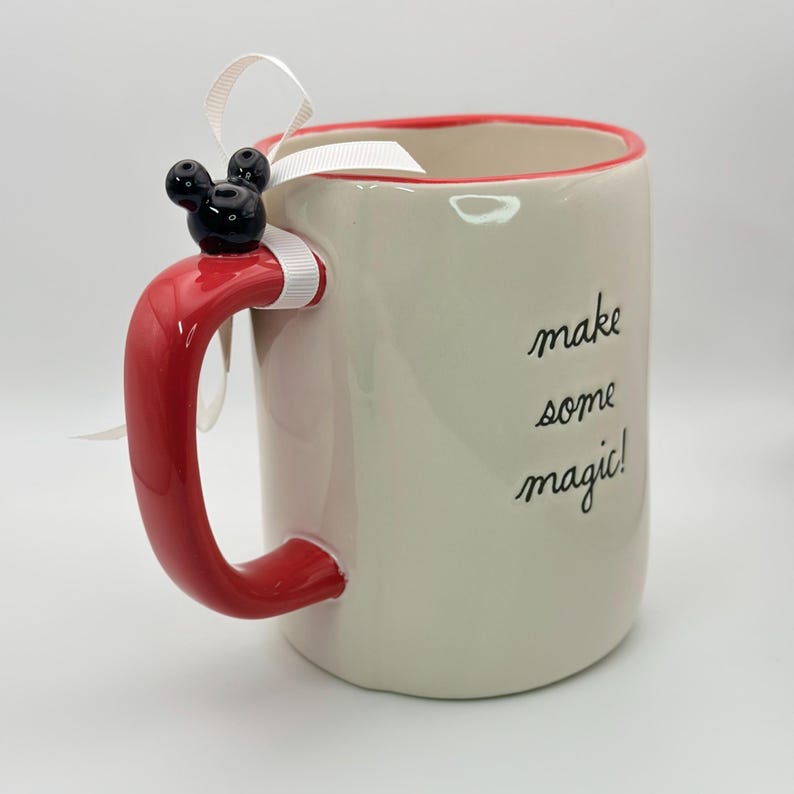 Op de afbeelding: Witte keramische mok met een rode handgreep en een rode rand. De mok heeft een zwart Mickey Mouse hoofd op de handgreep en de tekst "make some magic!" aan de zijkant.