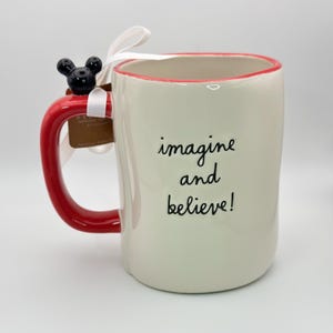 Op de afbeelding: Witte keramische mok met een rode rand en handvat. De mok heeft een zwart Mickey Mouse hoofd bevestigd aan het handvat en de woorden "imagine and believe!" gedrukt op de voorkant.
