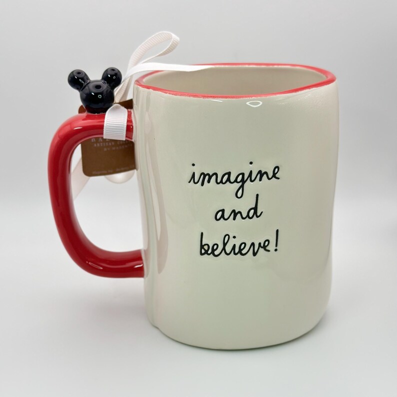 Op de afbeelding: Witte keramische mok met een rode rand en handvat. De mok heeft een zwart Mickey Mouse hoofd bevestigd aan het handvat en de woorden "imagine and believe!" gedrukt op de voorkant.