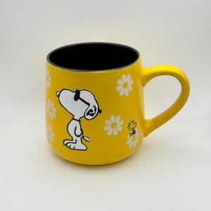 May include: A yellow ceramic mug featuring Snoopy wearing sunglasses and white daisy accents. The mug has a black interior and a yellow handle. A smaller image of Woodstock is also visible on the mug.