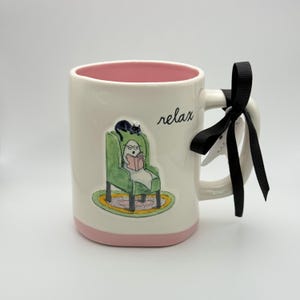 May include: White ceramic mug with a pink interior and base, featuring a whimsical illustration of a figure reading in a green armchair, a black cat, and the word "relax". A black ribbon is tied to the handle.