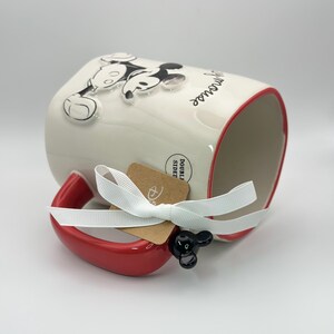 Op de afbeelding: Witte keramische mok met een rode rand en handvat. De mok heeft een zwart-witte afbeelding van Mickey Mouse met de tekst "Have a magical day". De mok heeft een zwart Mickey Mouse-oorhandvat en een bruin label met het Disney-logo en de tekst "Double Sided".