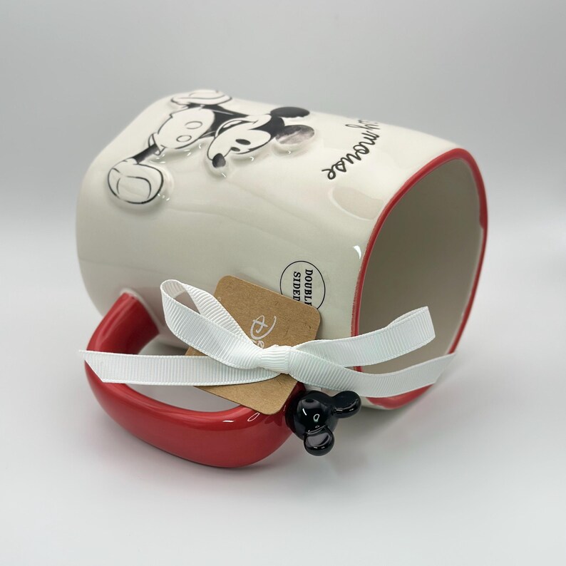 Op de afbeelding: Witte keramische mok met een rode rand en handvat. De mok heeft een zwart-witte afbeelding van Mickey Mouse met de tekst "Have a magical day". De mok heeft een zwart Mickey Mouse-oorhandvat en een bruin label met het Disney-logo en de tekst "Double Sided".