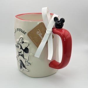 Op de afbeelding: Witte keramische mok met een rode rand en handvat. De mok heeft een zwart-witte afbeelding van Minnie Mouse met de tekst "Minnie Mouse" aan de zijkant. Het handvat heeft een zwart 3D Mickey Mouse-oor bovenop. Een bruin label met het woord "Disney" is aan de mok bevestigd met een wit lint.