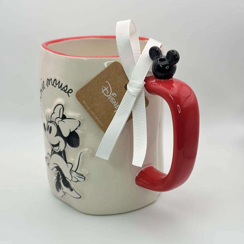 Op de afbeelding: Witte keramische mok met een rode rand en handvat. De mok heeft een zwart-witte afbeelding van Minnie Mouse met de tekst "Minnie Mouse" aan de zijkant. Het handvat heeft een zwart 3D Mickey Mouse-oor bovenop. Een bruin label met het woord "Disney" is aan de mok bevestigd met een wit lint.