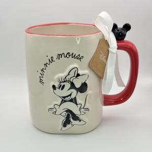 Op de afbeelding: Een witte keramische mok met een rode rand en handvat. Het heeft een zwart-witte afbeelding van Minnie Mouse met de tekst "minnie mouse" aan de zijkant. De mok heeft een klein zwart Minnie Mouse-oor aan het handvat en een bruin Disney-label.