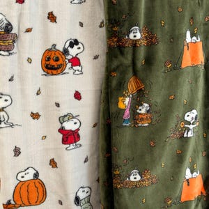 May include: Two pieces of fabric featuring Peanuts characters. The left side is cream-colored with Snoopy, pumpkins, and autumn leaves. The right side is olive green with Snoopy, Charlie Brown, and Woodstock playing in piles of fall leaves.