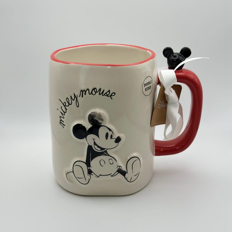 Op de afbeelding: Witte keramische mok met een rode rand en handvat. De mok heeft een zwart-wit afbeelding van Mickey Mouse met de tekst "mickey mouse" in zwarte schrijfletters. De mok heeft een dubbelzijdig ontwerp.