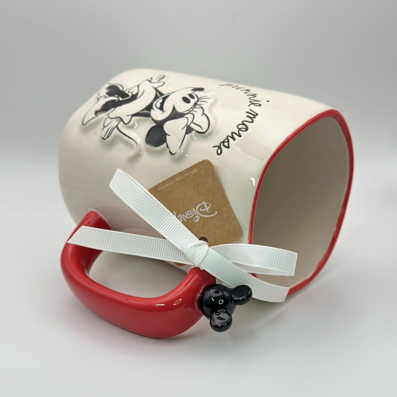 Op de afbeelding: Een witte keramische mok met een rode rand en handvat. De mok heeft een 3D-afbeelding van Mickey Mouse en Minnie Mouse met de tekst "you're the mouse that got away". De mok heeft een zwart Mickey Mouse-oorhandvat en een wit lint om het handvat geknoopt.