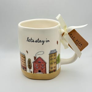 May include: A cream-colored ceramic mug featuring the black handwritten text "let's stay in" above a colorful illustration of houses and trees. A cream ribbon is tied to the handle with a brown rectangular tag labeled "RAE DUNN."