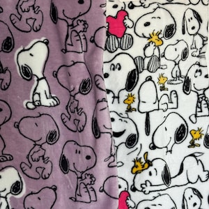 May include: Two plush fabrics, one lavender and one white, are shown. The lavender fabric features a repeating pattern of Snoopy in various poses. The white fabric has a similar pattern, with Snoopy and Woodstock. The Snoopy characters are in black and white.