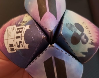 Può includere: Un gioco di indovinelli di carta colorato con le parole "BTS", "Wings", "Army" e "13:57" su ogni lato. Il gioco di indovinelli è piegato a forma di stella.