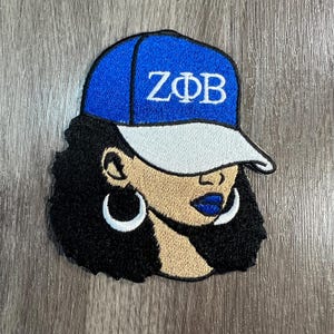 May include: Embroidered patch featuring a woman wearing a blue baseball cap with the Greek letters ZΦB in white. The woman has dark curly hair, hoop earrings, and blue lipstick. The patch is on a gray wooden surface.